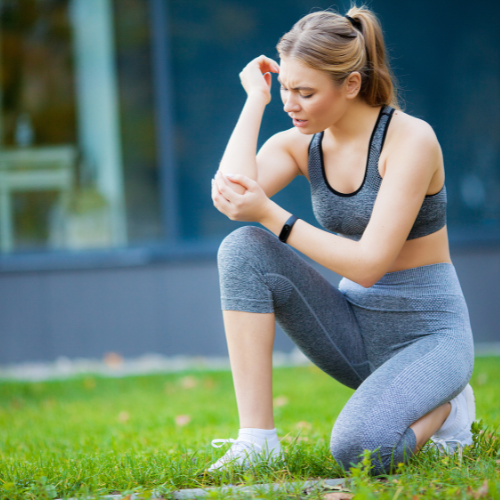 Woman in sport clothes outside kneeing down holding elbow in pain